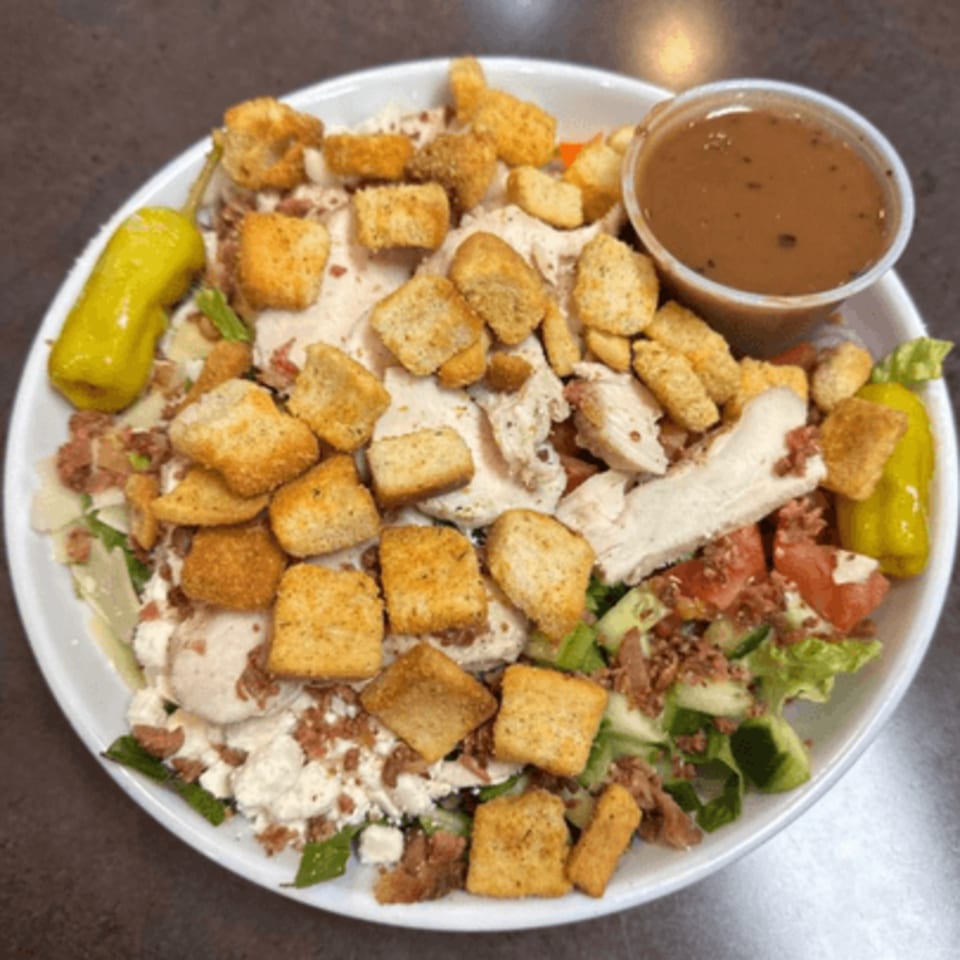 A large salad with chicken, croutons, bacon bits, and a side of dressing in a white bowl.