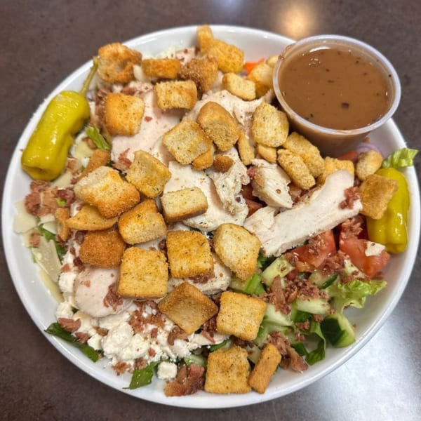 A large salad with chicken, croutons, bacon bits, cheese, and vegetables, with a side of dressing.